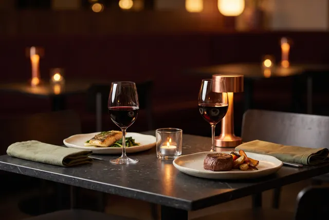 A table for two in a dim restaurant: stoneware plates with fish and steak, two glasses of red wine, olive napkins and a candle
